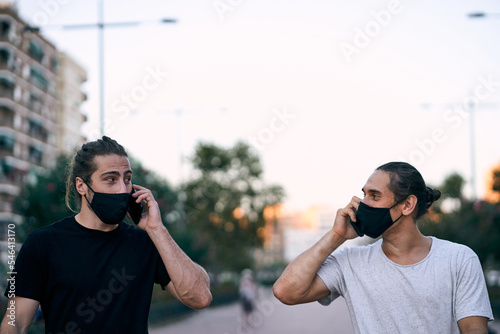 Fototapeta two caucasian young men walking on the city boulevard wearing face mask talking