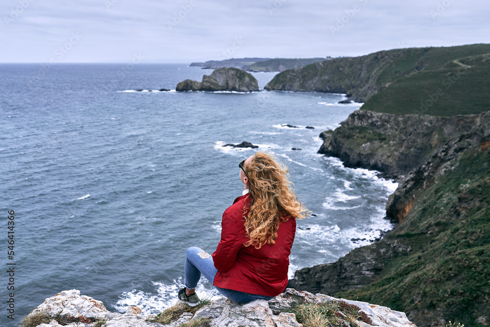 blond caucasian girl with red jacket and blue pants sneakers sitting on a rock looking at the sky admiring the beauty of the sea waves, cabo de penas, spain