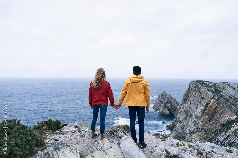 latin boy and caucasian girl in red and yellow jacket blue pants and sneakers standing holding hands looking at the horizon, cabo de penas, spain