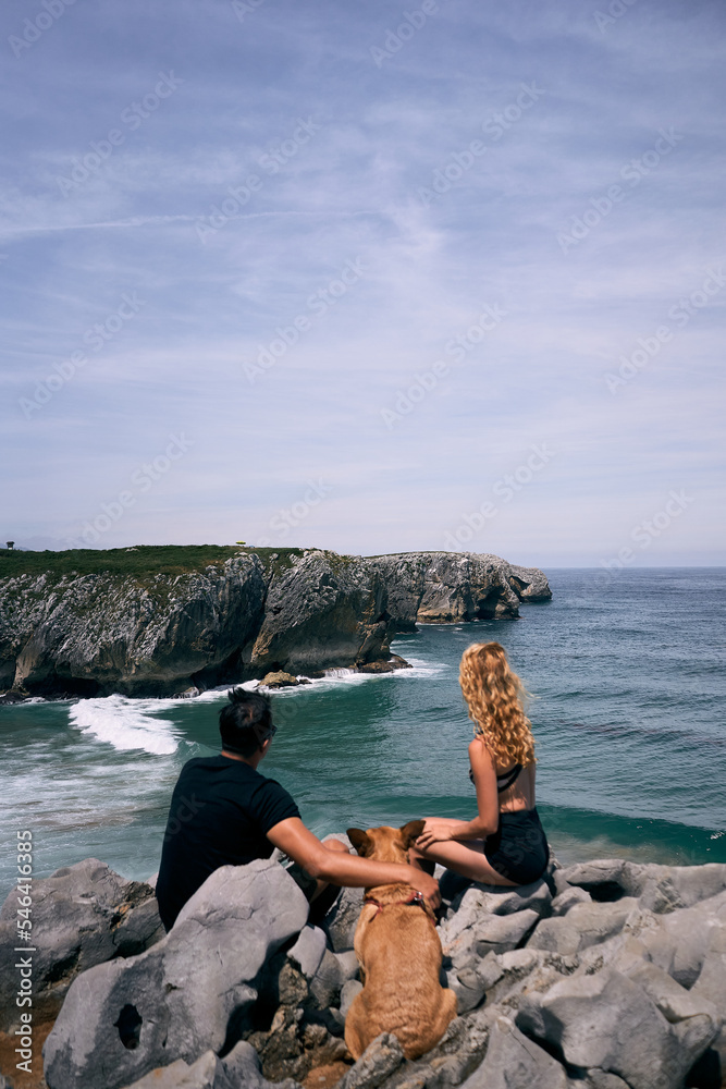 latin boy and blonde caucasian girl calm and relaxed with their dog contemplating the waves of the sea, ribadesella asturias, spain