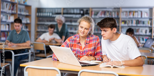 Positive female and male students teenagers study together at laptop while sitting in the school library