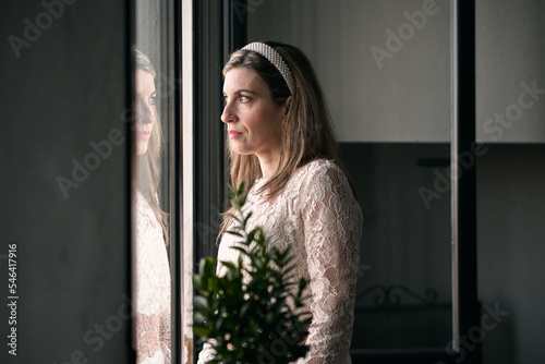 caucasian mature woman in white dress and headband in her hair standing looking out the window calm and relaxed next to a plant