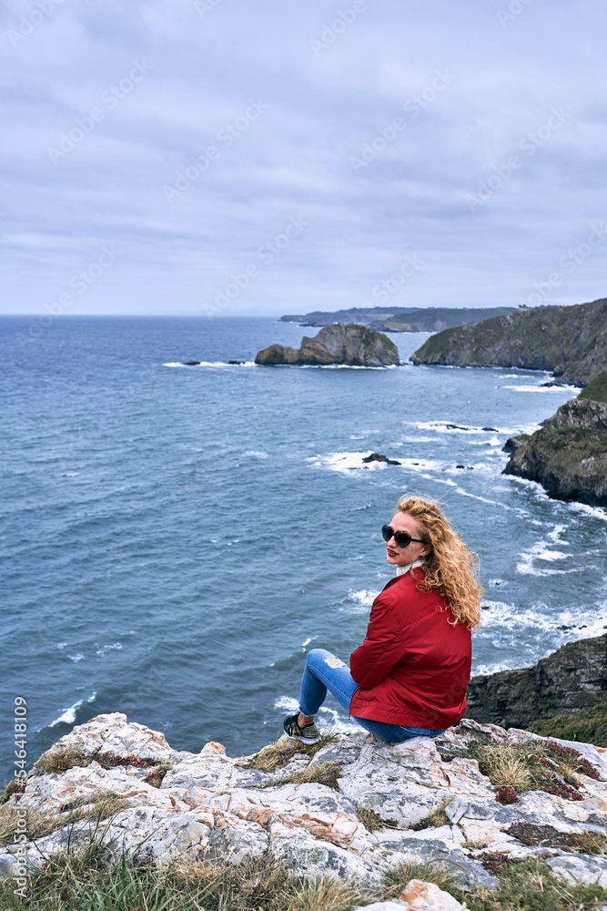 blonde caucasian young woman with red jacket and blue pants sneakers sunglasses sitting on a rock looking at camera next to the cliffs and sea waves, cabo de penas, spain
