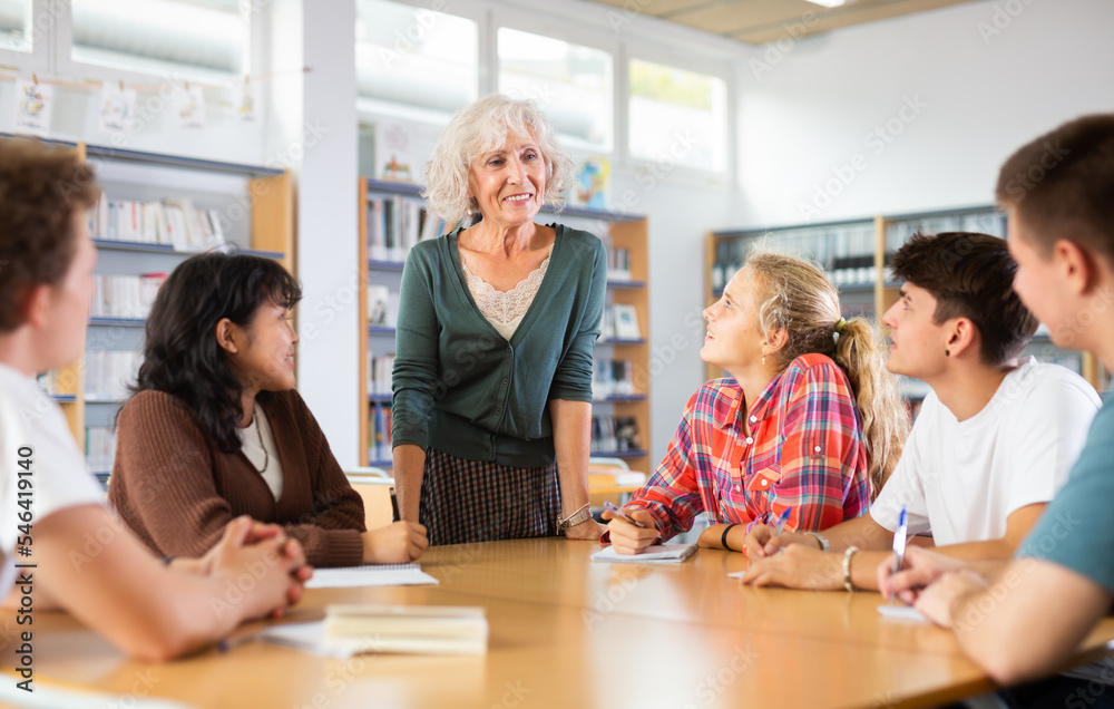 Obraz premium Group of fifteen-year-old schoolchildren with a mature female teacher are preparing for classes in the school library, discussing something