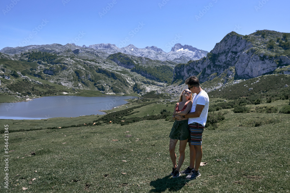 latin boy and caucasian girl hugging and looking at each other tenderly on the grass by the lake and mountains, covadonga asturias, spain