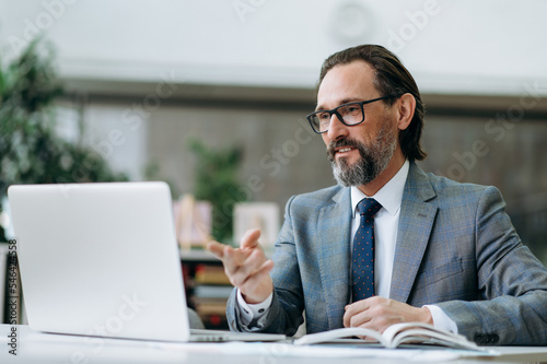 Elegant senior businessman is having a conversation on video conference. Concentrated male ceo communicate with colleagues, discussing important project