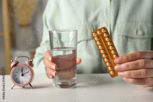 Woman holding blister of oral contraceptive pills and glass with water at white table, closeup