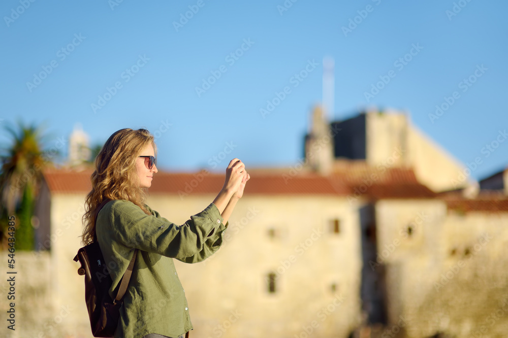 Charming young tourist woman taking photo using smartphone in old town of Budva, Montenegro. Attractive red-haired girl admiring of ancient town at sunset.