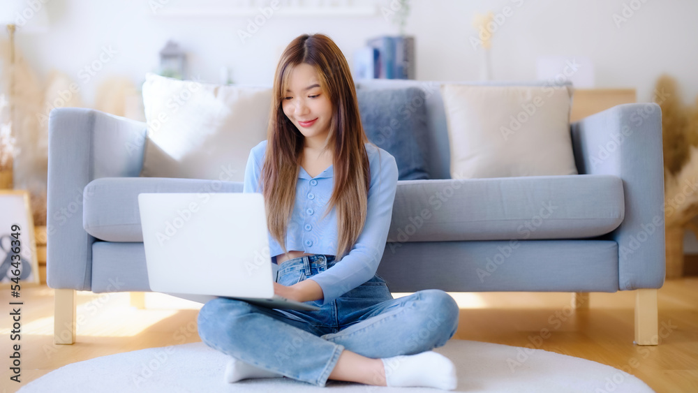 Young asian woman in good spirits working on laptop at home while sitting on the floor close to the couch Young asian woman in good spirits working on laptop at home while sitting on the floor close to the couch