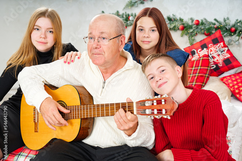 Grandfather playing guitar during christmas for grandchildren. Happy man 60s playing guitar christmas songs. Happy christmas atmosphere. © Ilja