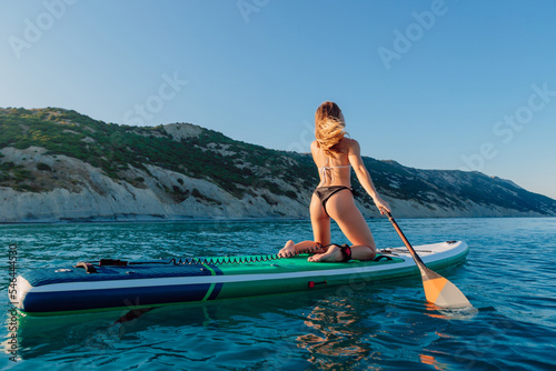 Sporty girl rowing on stand up paddle board at quiet sea. Attractive woman on SUP board in ocean with mountain coastline. © artifirsov