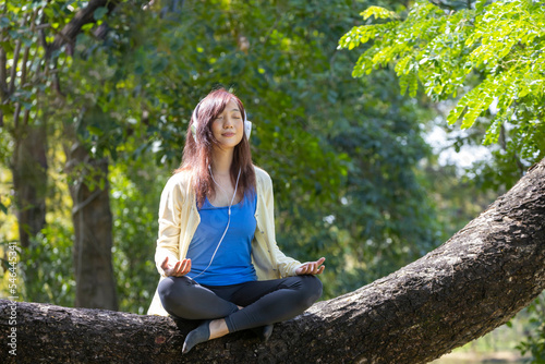 Young Asian woman meditating while sitting on the mature tree branch listening to the sound of nature from her mobile phone in the forest for yoga and inner peace meditation practice concept