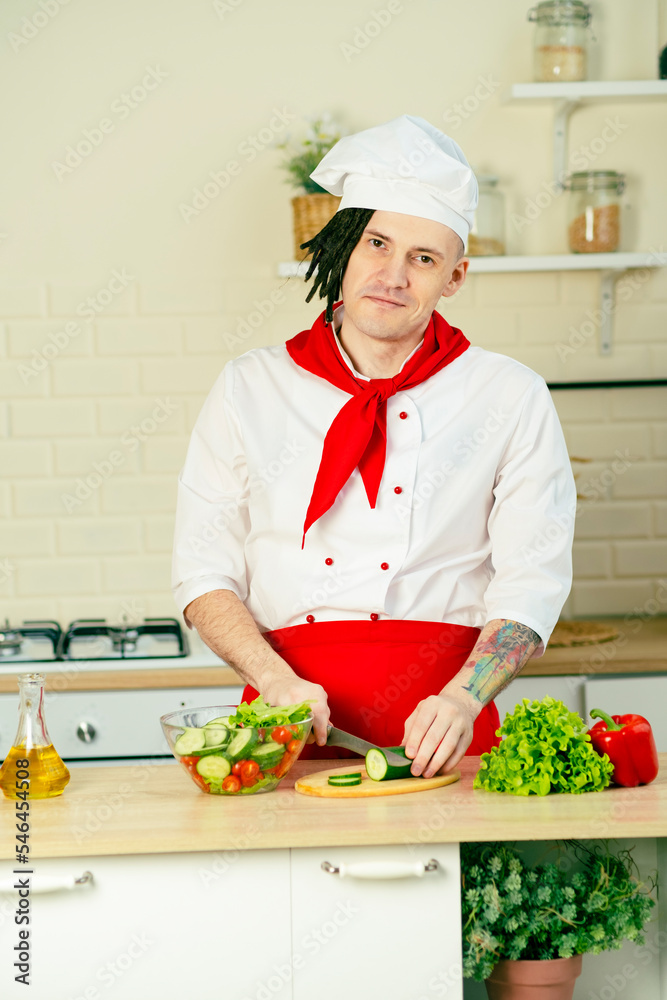 Young handsome chef cutting green cucumber on wooden board in kitchen. Positive guy with dreadlocks in uniform preparing vegetable salad. Young handsome chef cutting green cucumber on wooden board in kitchen. Positive guy with dreadlocks in uniform preparing vegetable salad.