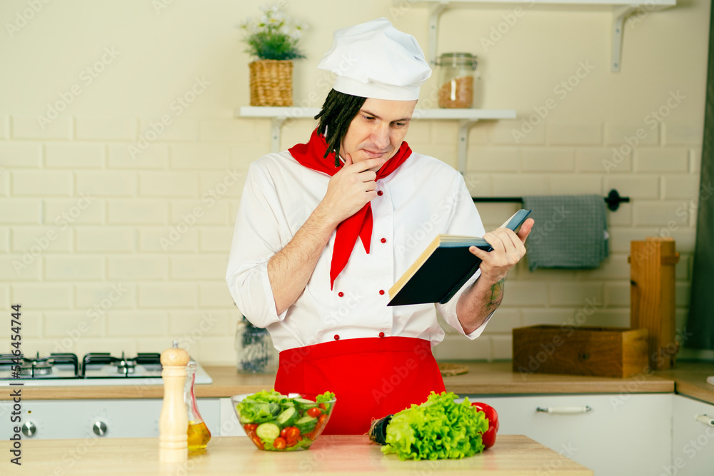 Young man dressed as chef reading recipe book in kitchen. Concept of food, cooking, recipes. Young man dressed as chef reading recipe book in kitchen. Concept of food, cooking, recipes.