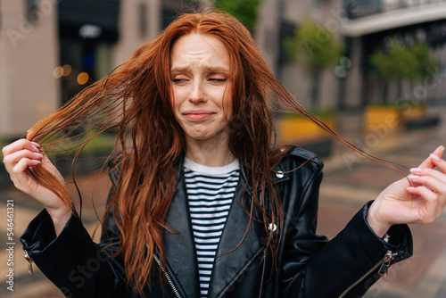 Portrait of sad attractive redhead young woman touching wet hair after autumn rain standing on beautiful city street. Front view of unhappy lady untangling hair after being caught in rain.