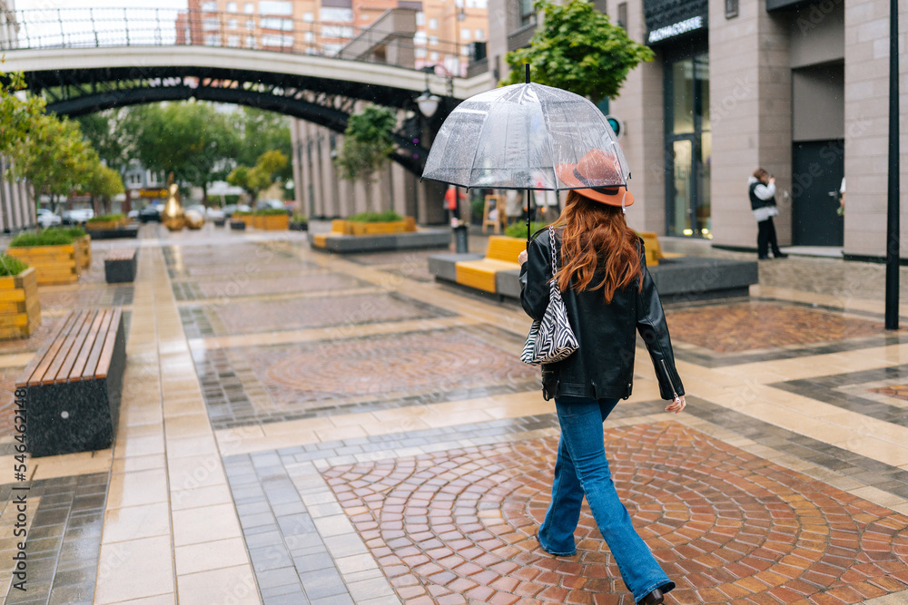Rear view to unrecognizable young woman in fashion hat walking on beautiful city street with transparent umbrella enjoying rainy weather outdoors. Concept of modern female lifestyle at autumn season. Rear view to unrecognizable young woman in fashion hat walking on beautiful city street with transparent umbrella enjoying rainy weather outdoors. Concept of modern female lifestyle at autumn season.