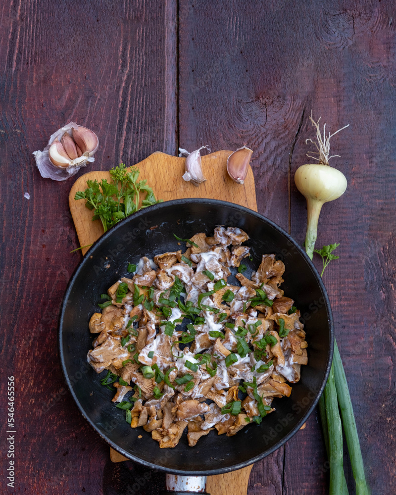 French-fried chanterelle mushrooms with onions and sour cream in a frying pan on a dark wooden background, garlic, onion and parsley and dill greens are laid out next to each other.