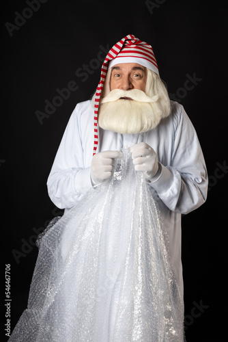 Santa Claus in pajamas and a cap squinted strongly, relaxing, shaking bubble wrap in his hands from excitement. The concept of a cool Santa with a squinted face for Christmas, only Black background.