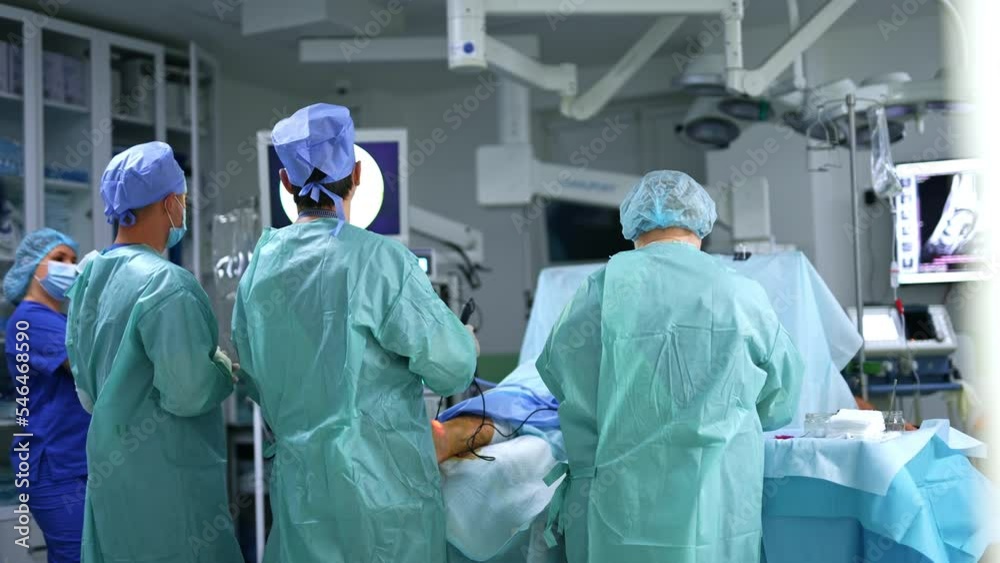 Group of surgeons working in modern surgery room. Chief doctor using device on the operated leg and watching the monitor. Group of surgeons working in modern surgery room. Chief doctor using device on the operated leg and watching the monitor.