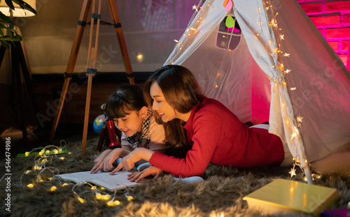 Happy Asian mother, mom and daugter reading a book together in a camp tent at home or house at night. Family people activity lifestyle. Kid photo