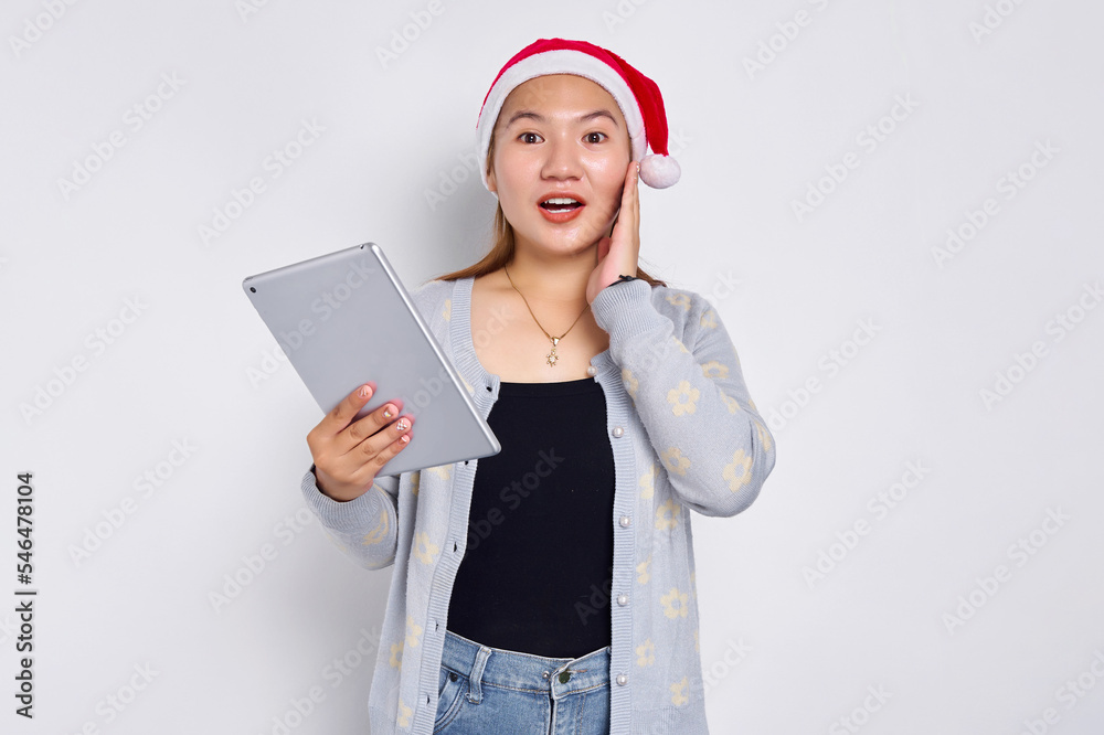 Excited young Asian woman in a hat Christmas using a digital tablet and touching her cheek isolated over white background. Indonesian people celebrate Christmas concept Excited young Asian woman in a hat Christmas using a digital tablet and touching her cheek isolated over white background. Indonesian people celebrate Christmas concept