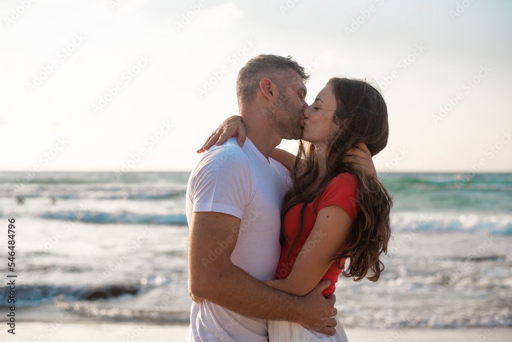 Happy couple hugs and kisses on beach on background clear sky and blue sea. Romantic date on seaside. Man in white t-shirt and woman in red t-shirt. Love and care, vacation and happiness. Happy couple hugs and kisses on beach on background clear sky and blue sea. Romantic date on seaside. Man in white t-shirt and woman in red t-shirt. Love and care, vacation and happiness.