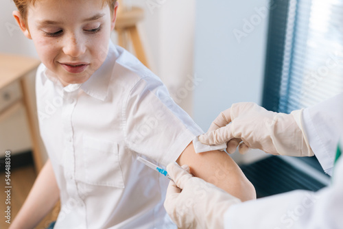 Close-up cropped shot of unrecognizable female doctor applying plaster on smiling child boy after vaccination injection at hospital. Concept of vaccination program, prevention of infectious diseases.