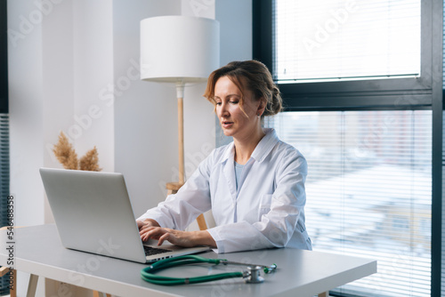 Portrait of blonde female practitioner wearing white coat working typing on laptop computer looking on display screen, sitting at table in light office room at hospital on background of window.