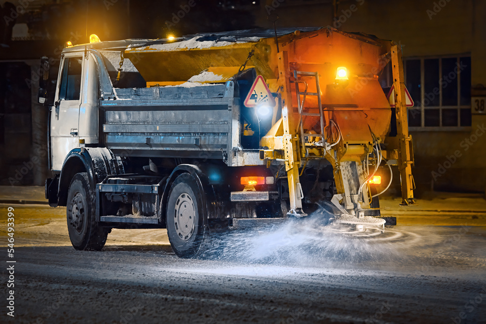 Truck spread salt and sand on snowy city road during blizzard at night shift, night work road maintenance. Snow and ice removing service. Salt spreading on snowy road, melt snow and ice. Deicing work