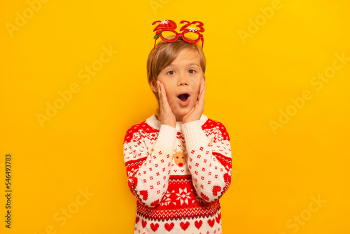 Surprised shocked little kid boy in Christmas winter sweater with party glasses 2023 keeps hands on cheeks and looking at camera on a yellow background.Happy New Year and Xmas celebration concept.