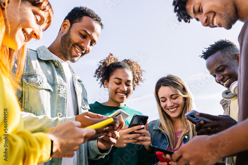 Young group of happy friends using smart mobile phone together outdoors. Multiracial teenage people having fun together sharing social media content on cellphone app. Technology lifestyle concept