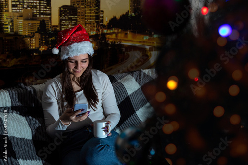 Young woman in santa hat smiles while looking at her phone and holds mug. Selective focus. Images for articles about leisure, winter, communication. © Natali Mali