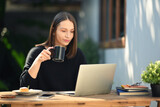 Beautiful young woman drinking hot coffee and working with laptop while sitting at the table in outdoor coffee shop