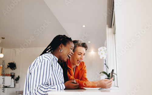 Happy business women laughing excitedly while using a smartphone together in a cafe photo