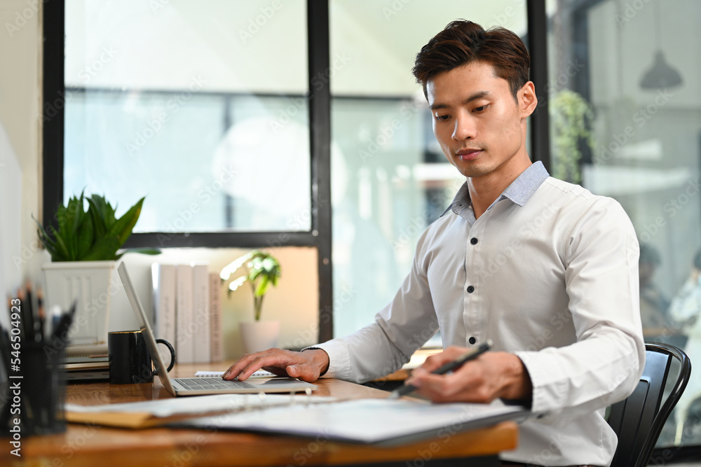 Professional male economist using laptop and financial document on wooden desk in modern office Professional male economist using laptop and financial document on wooden desk in modern office