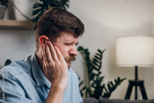 Close-up of upset young guy 30s touches sore ears suffering from sudden throbbing earache. Unhealthy caucasian male feeling unwell sitting on grey couch in living room at home. Tired ears concept