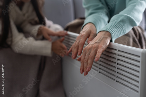 At the electric heater, people are warming themselves, covered with a warm blanket at home. hands of mother and daughter near the heater. Cold season and gas crisis in Europe