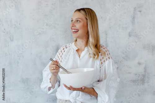 A happy woman whisking cream with a whisk in a white mixing bowl on a grey background.	 photo