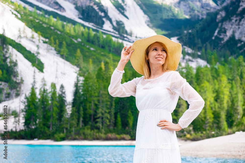 Smiling young woman on the Lake Braies in a white fluttering dress and hat on a sunny day 