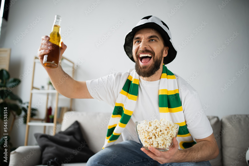 excited sports fan in hat and scarf holding bowl with popcorn and bottle of beer while watching championship. - obrazy, fototapety, plakaty 