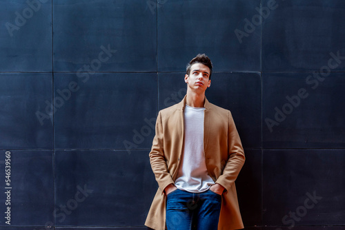 Young guy with modern hairstyle with coat, blue jeans and white sweeter leaning on outdoor wall photo