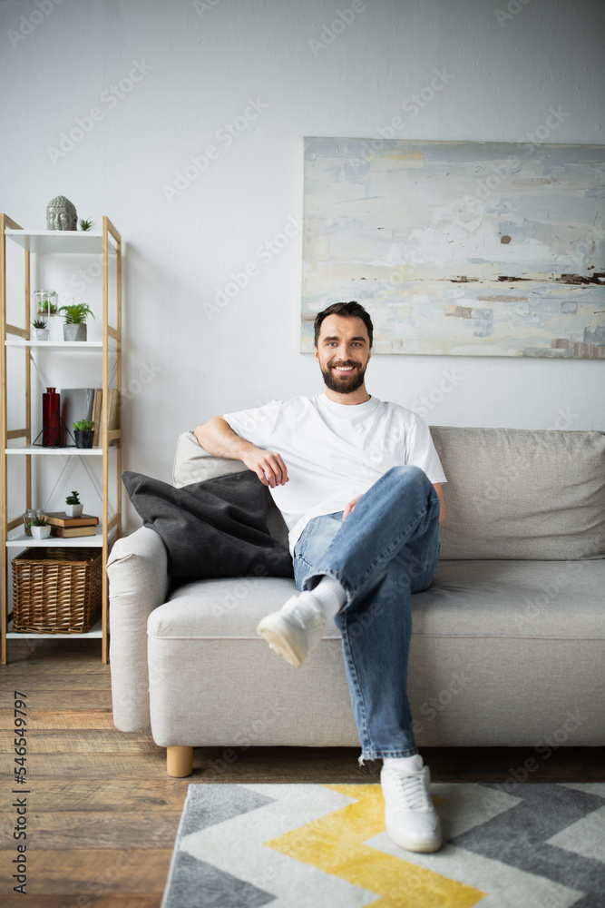 happy bearded man in jeans sitting on couch in modern living room.