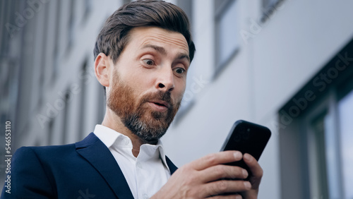 Close-up young surprised man looks at phone screen receiving notification reading good news rejoicing in success satisfied businessman gets excellent result from sales profitable offer salary increase photo