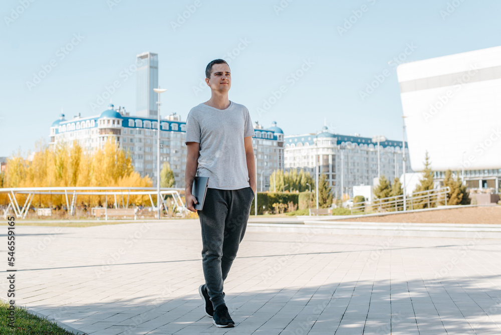 Young male freelancer or student in casual wear holding closed laptop while walking on street in city and looking away. Full length portrait of european guy in square outdoors Young male freelancer or student in casual wear holding closed laptop while walking on street in city and looking away. Full length portrait of european guy in square outdoors