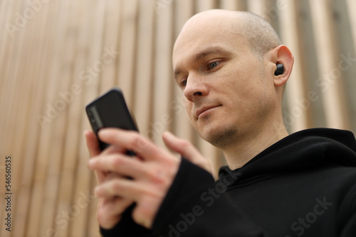 A young bald man with freckles. Guy in black hoodie using phone with wireless headphones and listen music.