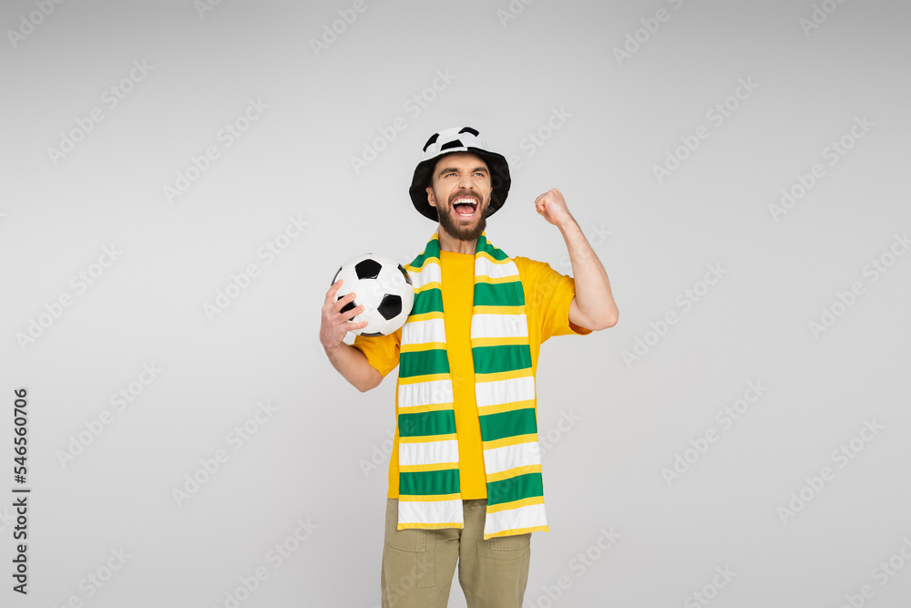 Fototapeta premium excited sports fan in hat and scarf holding soccer ball and showing triumph gesture isolated on grey.