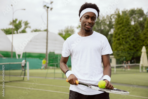Man of African appearance is playing tennis on tennis court and posing for photo. He is holding racket and ball. Guy wearing white t-shirt, headband and wristbands is looking at the camera and smiling © ABCreative