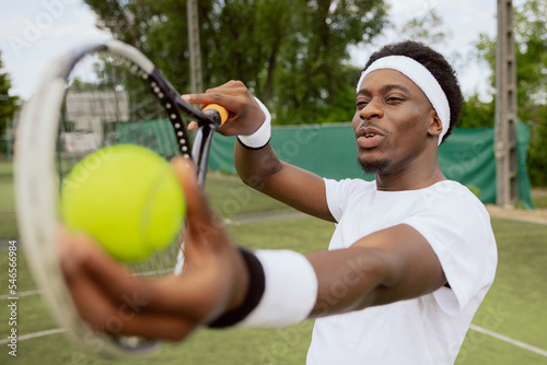 Close-up of tennis player of African appearance making serve. Man holds tennis ball and racket and prepares to hit ball. He is focused on hitting. Boy is wearing white T-shirt, bandage and wristbands.