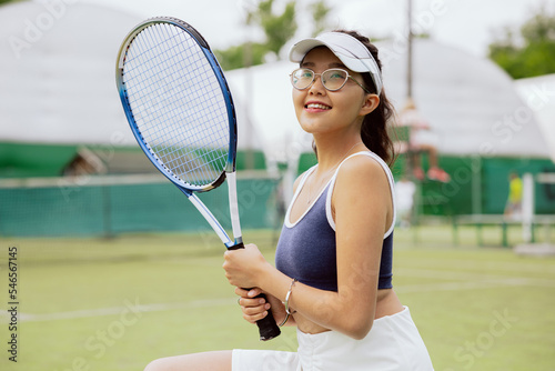After winning tennis match, young athlete takes photo. She smiles happily at the camera and holds racket in hands. The beauty is dressed in top and white skirt and has tennis cap on head.