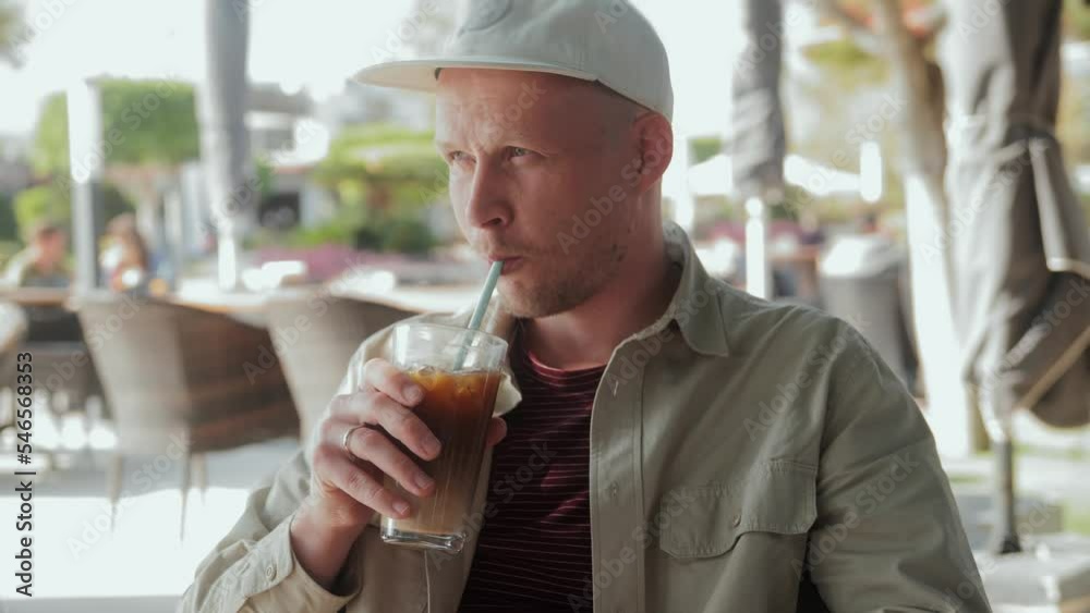 A young man is sitting in a cafe in the fresh air and drinking iced coffee. High quality 4k footage A young man is sitting in a cafe in the fresh air and drinking iced coffee. High quality 4k footage